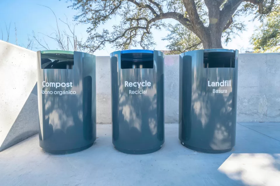 Three grey trash cans labeled with Compost, Recycle, and Landfill in English and Spanish on a concrete walkway in a park in Austin, Texas.